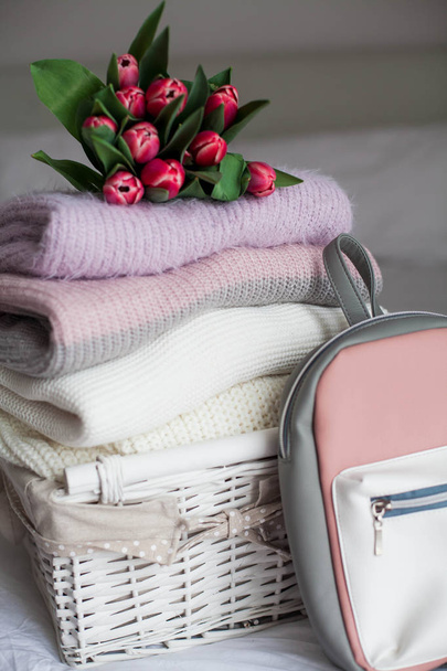 A basket with clean things on the bed. Cozy. Knitted sweaters, backpack, bouquet of tulips. Spring clothes. - Foto, afbeelding