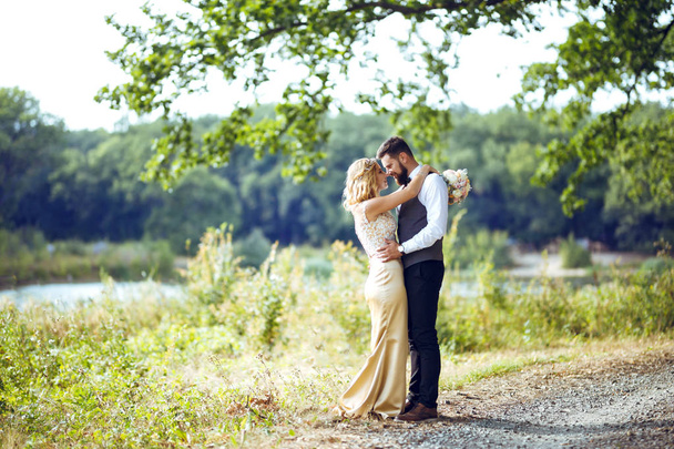 Stylish couple of happy newlyweds posing in the park on their wedding day. Perfect couple bride, groom posing and kissing  - Фото, изображение