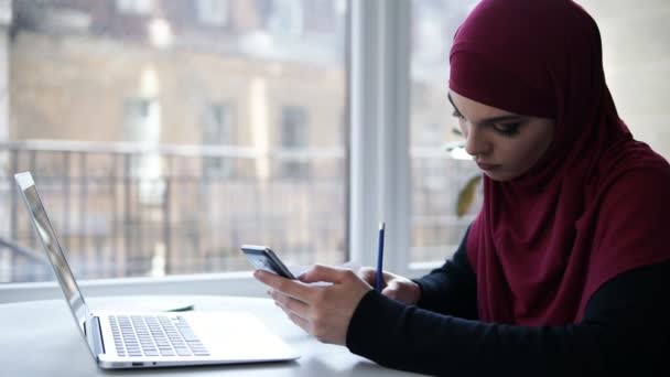 Young suposedly muslim girl with purple hijab on her head writes something down from her smartphone, sitting in a light indoor space with glass windows on the background - Materiał filmowy, wideo