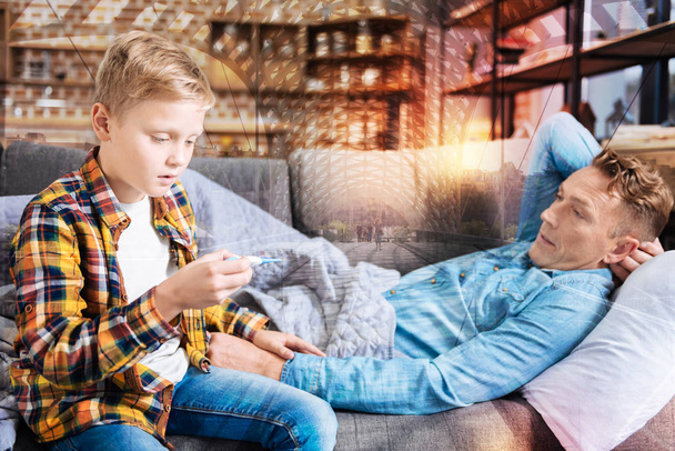 Astonished boy looking at the thermometer while sitting with his ill father - Фото, зображення