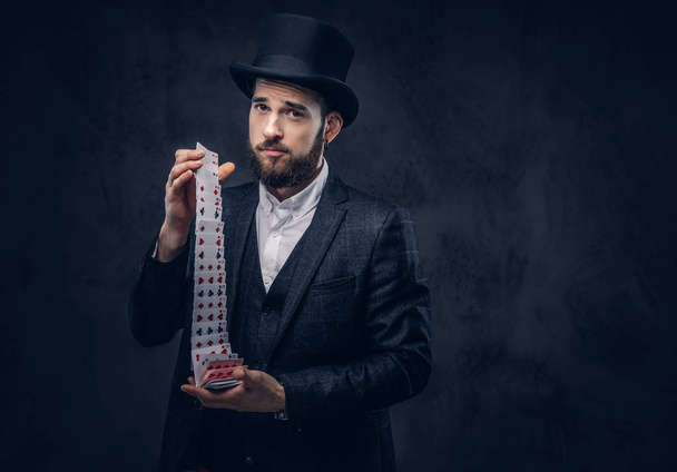 Retrato de un mago guapo en un palo negro, mostrando truco con jugar a las cartas sobre un fondo oscuro
. - Foto, Imagen