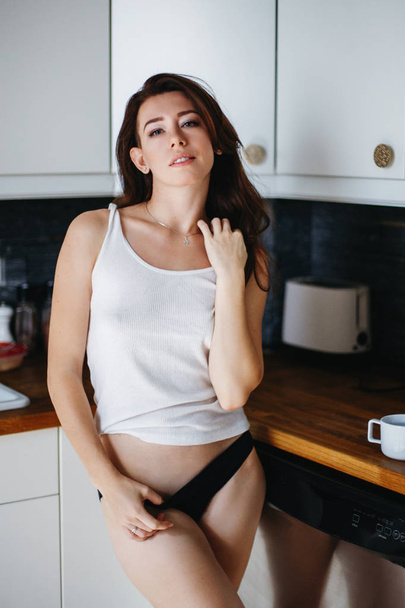 Porträt einer schönen jungen sexy Kaukasierin in weißem T-Shirt und schwarzem Höschen, die in der Küche steht. Natürliche Schönheit Jugendlook. Mädchen in Unterwäsche zu Hause - Foto, Bild