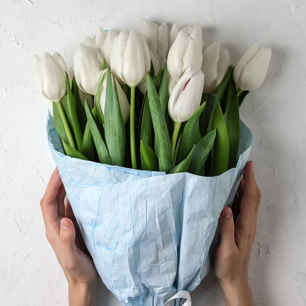 A bouquet of white tulips in blue wrapping paper with woman hands on a white concrete background. Top view. Flat lay. Postcard for Easter, Mothers Days. A square size for social networks. - Fotó, kép
