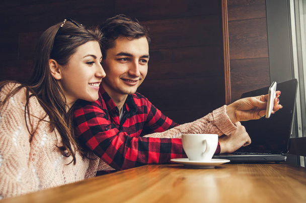 Νεαρό ζευγάρι παίρνει selfie και απολαμβάνει καφέ στο καφέ ενώ χρησιμοποιώντας ένα φορητό υπολογιστή - Φωτογραφία, εικόνα