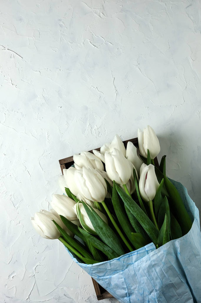 A bouquet of white tulips in blue wrapping paper in wooden box on a white concrete background. Top view. Flat lay. Postcard for Easter, Mothers Day and Spring Holidays. - Fotó, kép