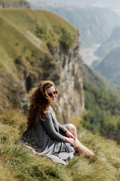 Young beautiful girl in a long dress is sitting on a cliff in the background of mountains - Foto, Bild