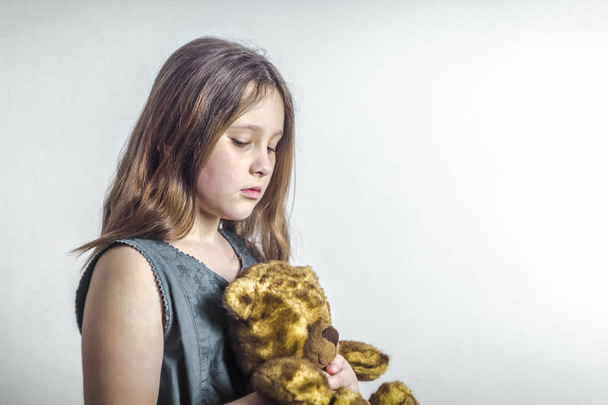 Девочка плачет, слеза течет по ее щеке, в руках плюшевого медведя, на белом фоне. Оскорбление
. - Фото, изображение