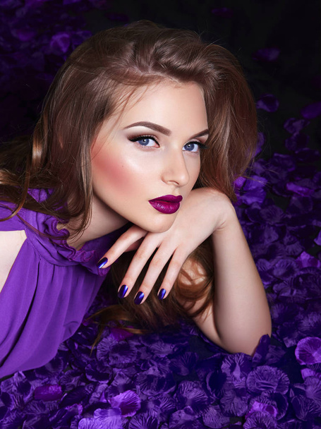 The girl in the petals. Beautiful young girl lies in the violet petals in a long dress. Glamor, luxe. Hair - curls. Makeup - arrows, purple lipstick. Love, romance. - Photo, Image