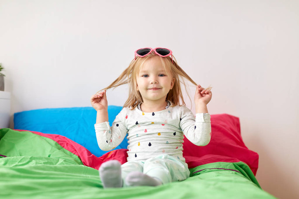 smiling little girl with sunglasses on bed at home - Фото, изображение