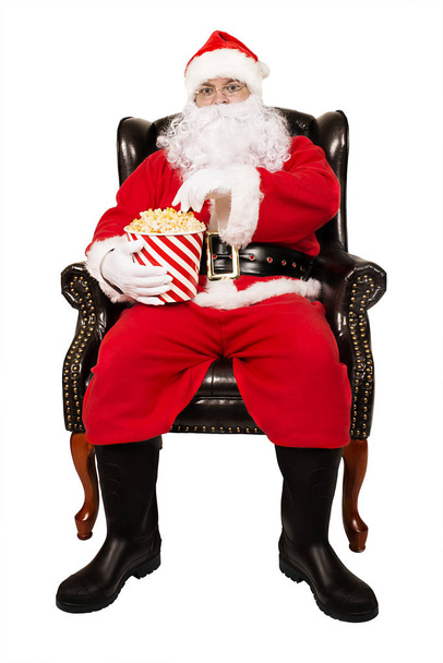 Santa Claus sits in chair is eating popcorn, isolated on white background. - Фото, зображення