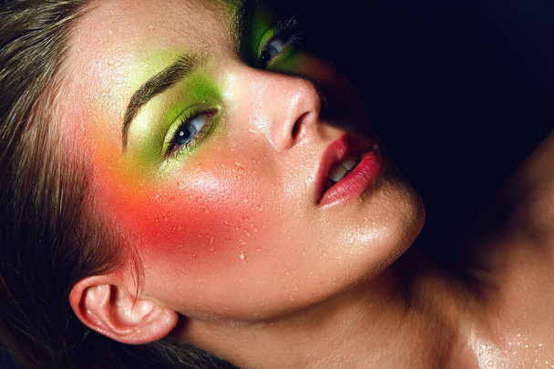 Beautiful young attractive girl lies. A large portrait is a face. Bright colorful make-up - green and pink colors. Drops of water on the skin, splashes, wet face. Cosmetics, healthy skin. - Fotografie, Obrázek