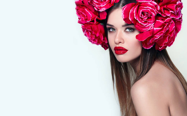 Beautiful girl with a big wreath on the head of a large red roses.Gentle make-up on the face of the girl. Red lipstick on the lips.Portrait of a girl with a crown of flowers in the studio. - 写真・画像