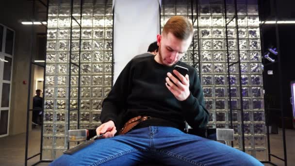 der Friseur der Mann sitzt auf einem Stuhl und benutzt das Smartphone, das auf den Kunden wartet - Filmmaterial, Video