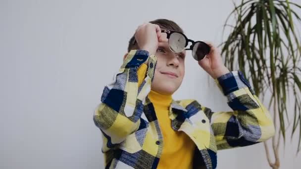 A handsome little boy with blue eyes wears sunglasses and looks aside. In the room, on a white background - Imágenes, Vídeo