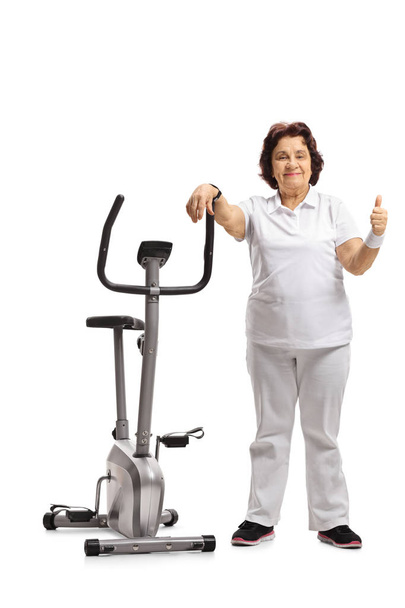 Full length portrait of an elderly woman leaning on an exercise bike and making a thumb up sign isolated on white background - Zdjęcie, obraz