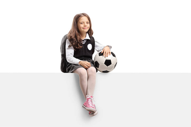 Cute little schoolgirl with a football sitting on a panel isolated on white background - Foto, Imagem