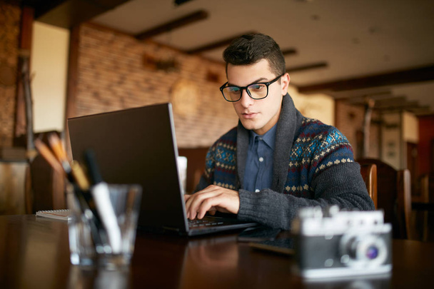 Fiatal, vonzó csípő férfi szemüveg és téli pulóver dolgozik a laptop a kávézóban. Szabadúszó üzletember, böngészés az interneten: coffeshop. Fotós, fotók retusálása. Vintage kamera asztalra. - Fotó, kép