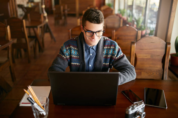 Lachende aantrekkelijke hipster man in glazen en winter trui werken op de laptop in café. Freelancer zakenman surfen op internet bij koffieshop. Fotograaf foto's retoucheren. Vintage camera op tafel. - Foto, afbeelding
