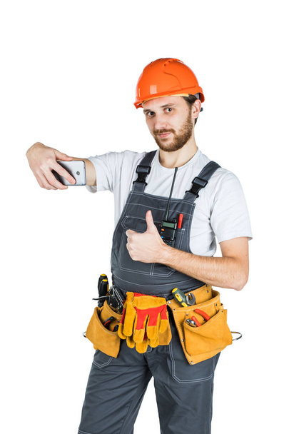 Bir işaret gösteren ve telefon resim alma genç inşaat işçisi gülümseyerek. Onarım ve inşaat. Beyaz arka plan üzerinde izole. - Fotoğraf, Görsel