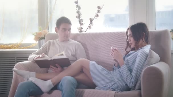 幸せな若い魅力的なカップルがソファに一緒に座って、男が本を読んで、少女は携帯電話を使用します。 - 映像、動画