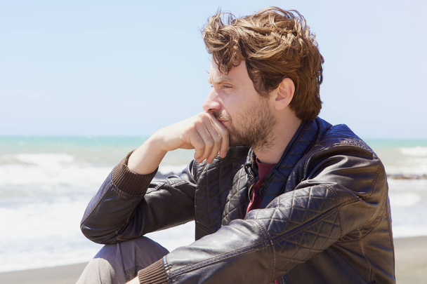 Красивый мужчина с голубыми глазами и бородой сидит перед океаном зимой глядя вид сбоку выстрел
 - Фото, изображение