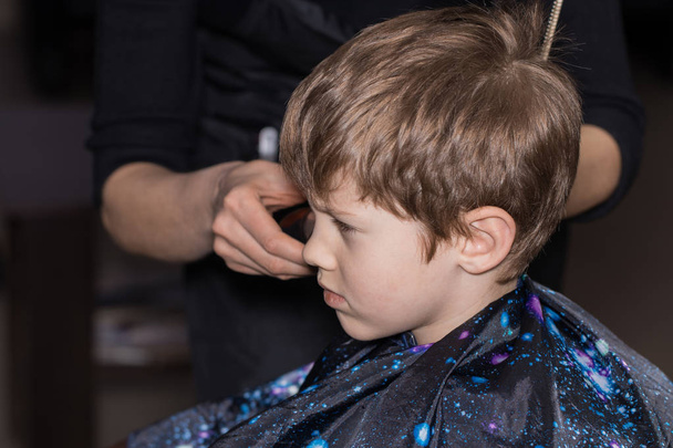 Вид сбоку на симпатичного мальчика, стригущегося парикмахером в футболке
 - Фото, изображение