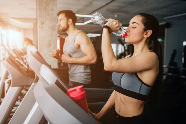 Beautiful and well-built girl is running on the running machine besides her sport partner and drinking water from the bottle while young man is just having workout - Photo, image