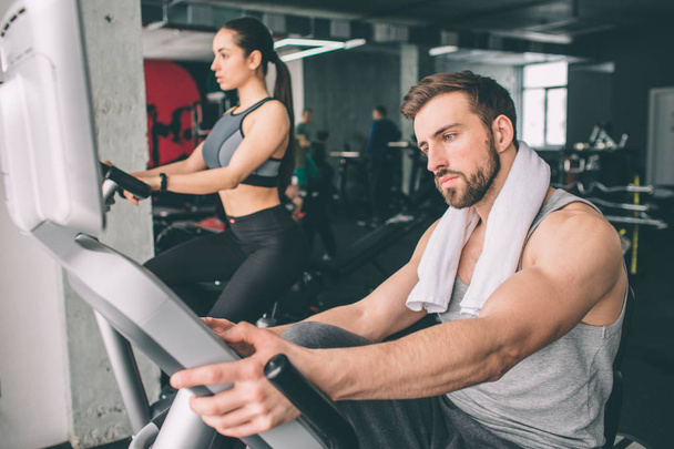 Close up of two young people training on the exercise bike. They look serious and concentrated on the workout. - Foto, afbeelding