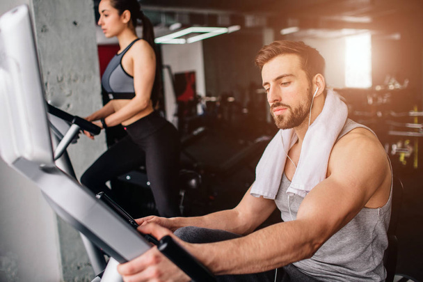 Close up of a guy wokring on the exercise bike and his girfriend doing the same thing further down. Both of them are serious and concentrated. - Фото, зображення