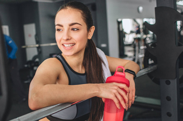 Close up of fantastic and gorgeous brunette that is resting after hard working exercises. She is laning on the fingerboard and holding pink bottle of water. She looks happy and satisfied. - Foto, afbeelding