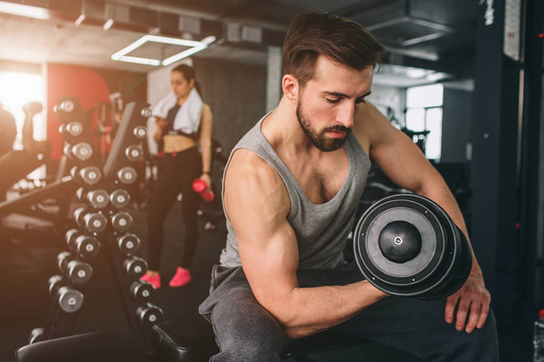 Close up of strong guy that exercising with a dumbbell on the sport bench while hir girlfriend is standing nearby, resting an looking to the phone. - Foto, Imagem