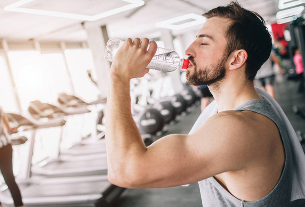 Close up of a handsome guy drinking water from the bottle and looking straight forward. He is having some rest after workout. Cut view - Foto, afbeelding