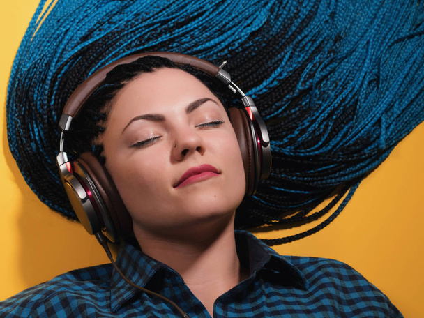Beautiful young girl with african blue braids is listening to music with headphones and dancing. Woman on bright yellow background. Dyed Hair moves. - Photo, image