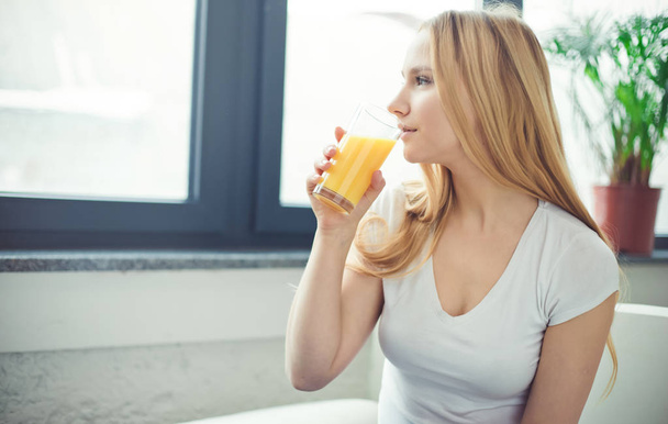 Smiling beautiful young blond woman sitting at home on the couch with a glass of orange fresh. - Foto, Imagem