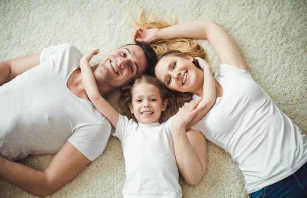 Top view of Young happy family. Beautiful and smiling father and mother with daughter lying on the floor at home and hugging looking at the camera. - 写真・画像