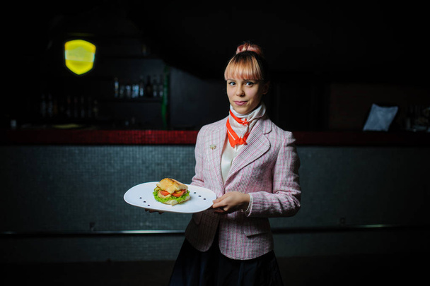 Nuori tarjoilija vaaleanpunaisine hiuksineen tarjoilee voileivän vintage-kahvilassa.
 - Valokuva, kuva