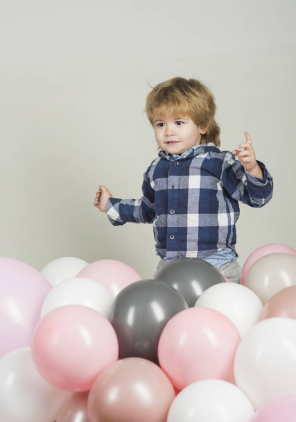 O menino salta em balões cor-de-rosa. O miúdo engraçado joga sozinho no fundo branco. Balões rosa, branco e cinza para celebração. Rapaz giro com camisa azul e calças de ganga cinzentas. Roupas para crianças
 - Foto, Imagem