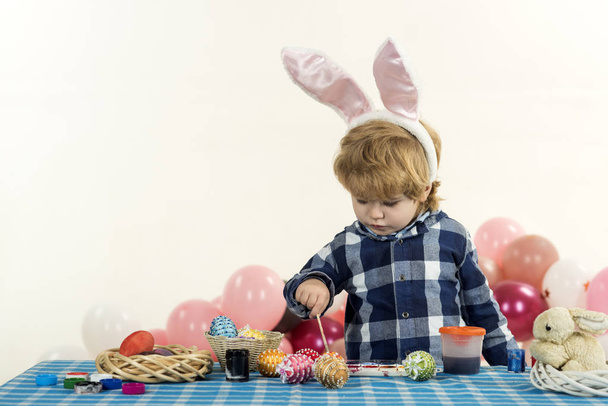 Chico dibuja huevos de Pascua en casa en la mesa en acogedora sala de estar. Tradiciones familiares y creatividad. Orejas rosadas de conejito en la cabeza del chico guapo. Preparación para la Pascua
 - Foto, imagen