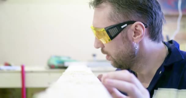 Ein Mann in einer Tischlerei mit Schutzbrille bläst Sägemehl vom Brett und betrachtet die Qualität des Holzes. - Filmmaterial, Video