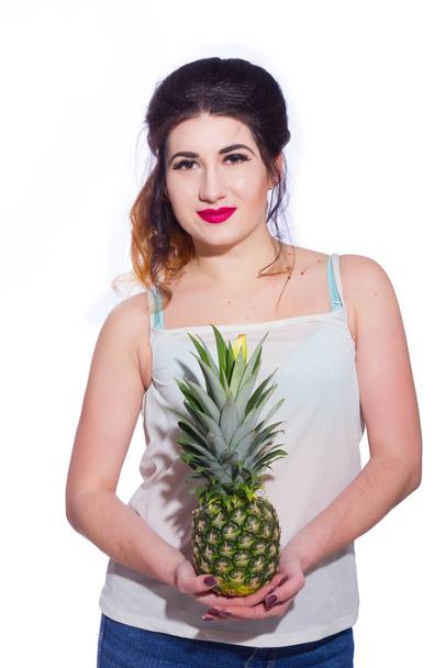 Summer studio portrait of a beautiful young brunette woman with long hair. A girl in a summer T-shirt and jeans is holding a pineapple in her hands. Concept: summer photography and a sunny mood - Foto, Imagem
