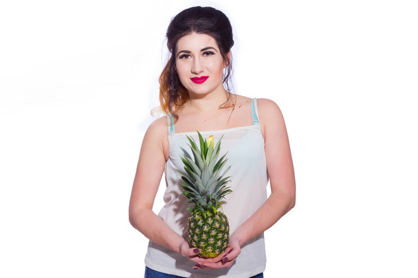 Summer studio portrait of a beautiful young brunette woman with long hair. A girl in a summer T-shirt and jeans is holding a pineapple in her hands. Concept: summer photography and a sunny mood - Фото, изображение