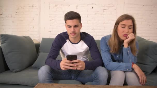 Schwenkbild eines jungen Mannes, der soziale Medien auf dem Handy nutzt und seine Freundin auf dem heimischen Sofa ignoriert, mit der frustrierten und gelangweilten Frau, die sich von ihrem Freund im Internetsuchtkonzept ignoriert fühlt - Filmmaterial, Video