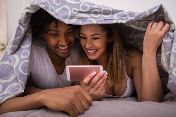 Bella coppia felice che gioca con il telefono nel letto sotto la copertura mentre sorride
 - Foto, immagini