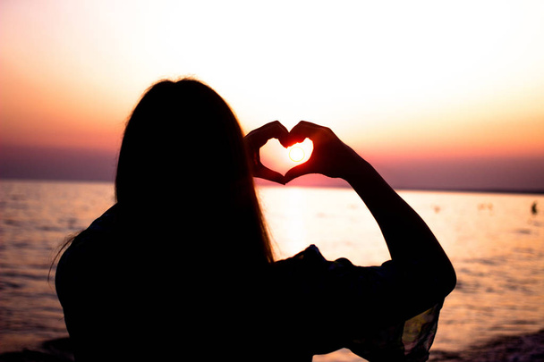 Silhouette of the heart from the hands of the symbol of love Young woman on the sea on sunset background - Photo, Image