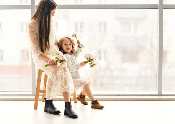 Mother and daughter have fun standing near a huge window, the daughter gives her mother flowers on her mother's day. - Foto, Bild