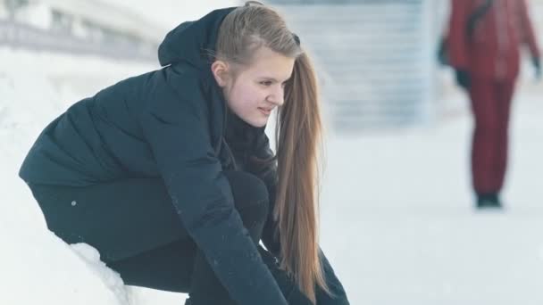 Εφηβική μακρυμάλλης κορίτσι που κάθεται στο χιόνι σφίγγοντας τα κορδόνια στα πατίνια και χαμογελαστός - Πλάνα, βίντεο