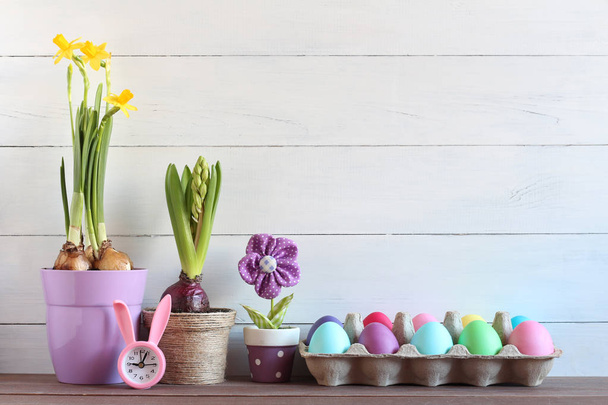 Easter still life. Color eggs, watch with ears of a rabbit, spring flowers in pots - Foto, Bild