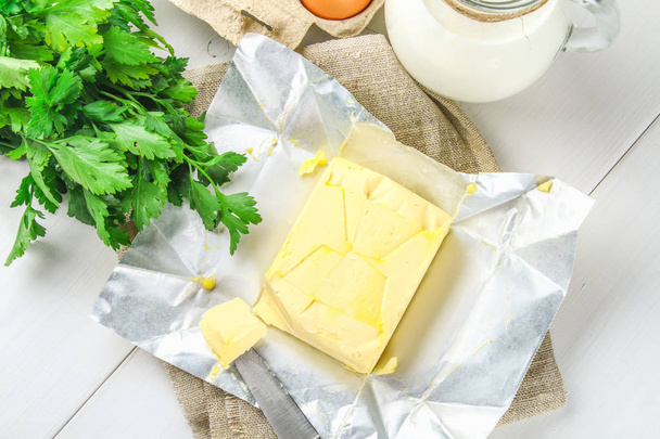 Ein Stück Butter wird auf einem Holzbrett mit einem Messer in Stücke geschnitten, umgeben von Milch, Eiern und Petersilie auf einem weißen Tisch. Zutaten zum Kochen. - Foto, Bild