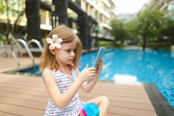 kleines Mädchen benutzt Tablet, das in der Nähe des Swimmingpools sitzt - Foto, Bild