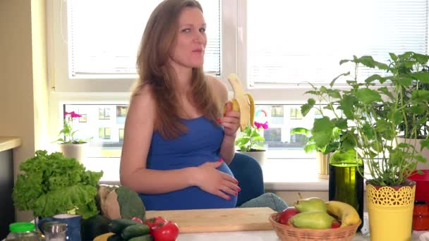 Beautiful caucasian pregnant woman eating banana in kitchen - Materiał filmowy, wideo